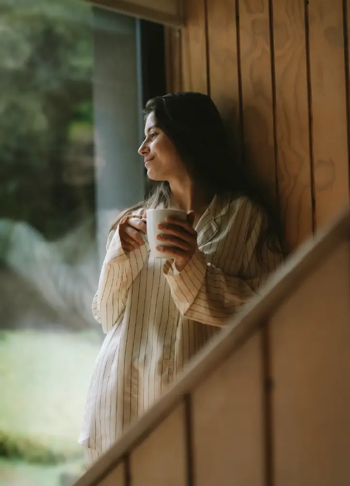 Mujer tomando café en el refugio, disfrutando del paisaje desde la ventana en un ambiente hogareño.