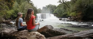 Dos mujeres sentadas en un tronco junto al río con una hermosa cascada y bosque en el Parque Nacional Puyehue