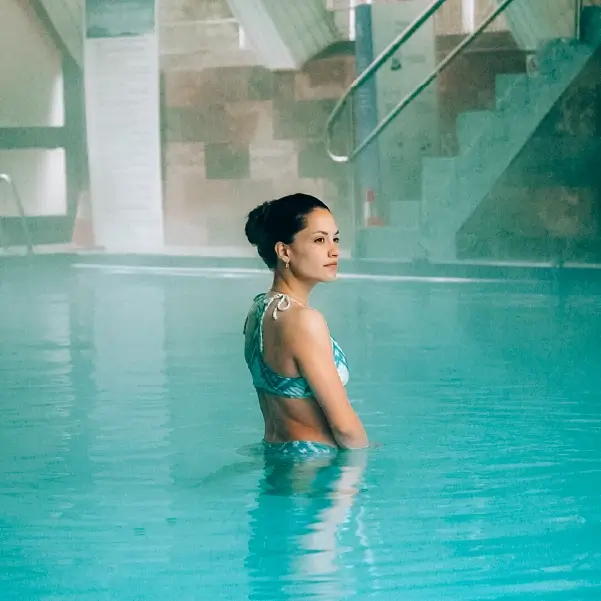 Mujer en piscina termal techada mirando al horizonte, reflejo celeste del agua