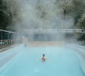 Mujer de espalda en la piscina abierta de Termas Aguas Calientes con bosque y vapor de fondo