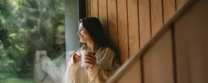 Mujer sonriendo con una taza de café en la ventana de un refugio de madera, con vegetación verde afuera